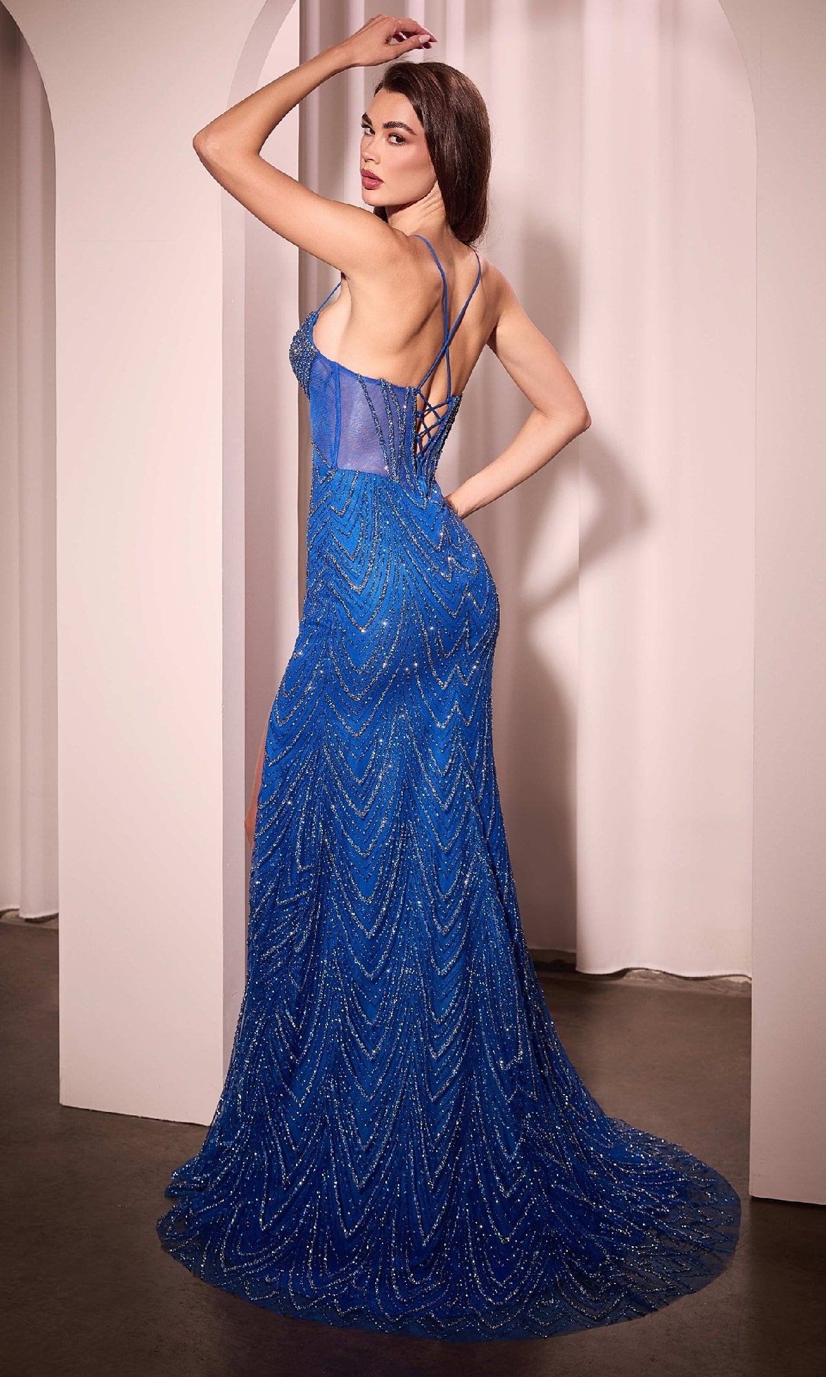 A woman poses in the Ladivine C163 Lace-Up Long Glitter Prom Dress, a sleeveless blue gown with sheer panels, beaded designs, and a lace-up back, standing before white arches.